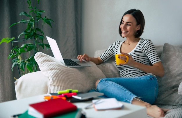 Close up photo of a gorgeous smiling woman working on a laptop while she lay on the big couch and drinking coffee at home