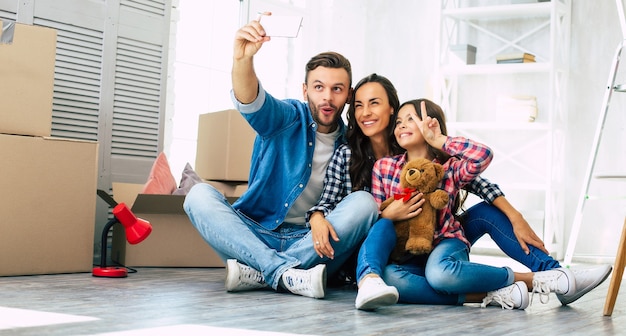 Close up photo, joyful family is taking selfie together in their new house in front of a pile of cardboard boxes