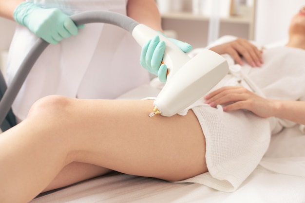 Close up photo of the legs of woman on the medical couch and professional specialist using a modern tool while removing vascular nets on the leg