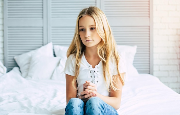 Close up photo of little sad blond girl is sitting on her bed in room