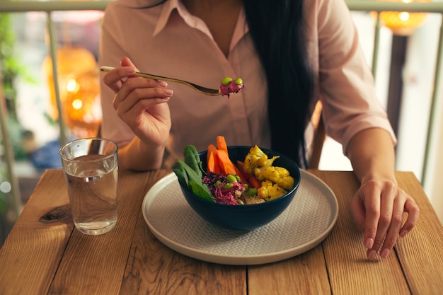 Close up photo of a long haired woman in a pink blouse sitting at the table with a glass of water and eating her vegetable meal with a fork