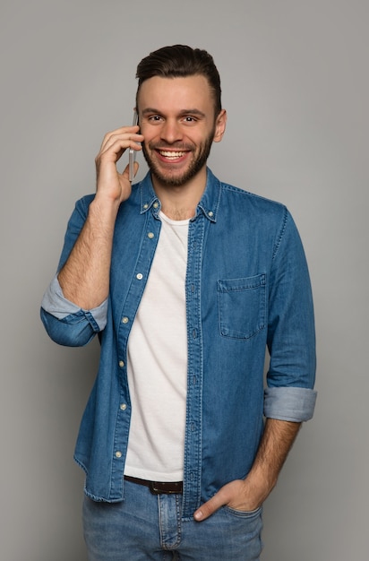 Close-up photo of a modern man in denim outfit, who is smiling, while talking on the phone and holding his left hand in a pocket.