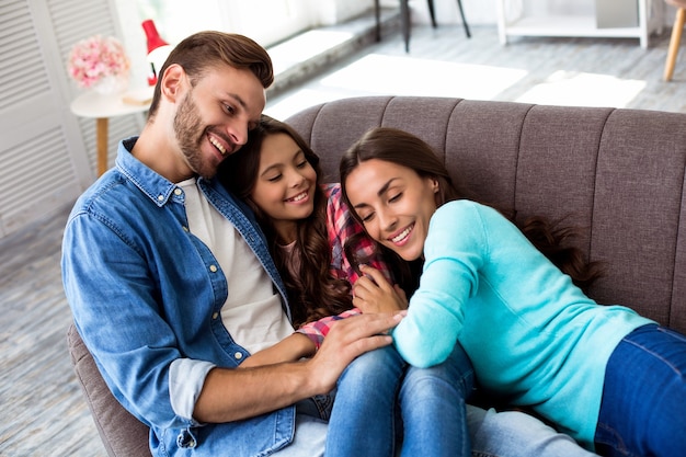 Close up photo of mother, father and their daughter posing together for a joint photo in their stylish living-room, hugging with radiant smiles