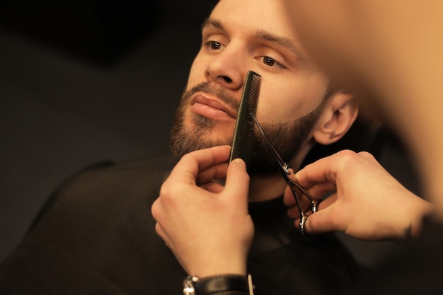 Close-up photo of a professional hairstylist, who is grooming beard of his young client, who is looking in the mirror during the procedure.