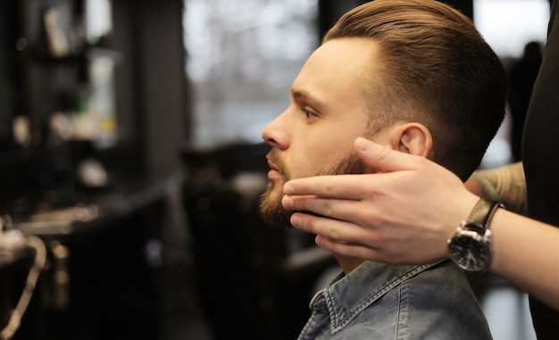 Close-up photo of a professional hairstylist, who is grooming beard of his young client, who is looking in the mirror during the procedure.