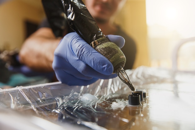 Close up photo of professional tattooer artist is gaining in the tattoo machine with black ink and going makes picture on the arm of a young man