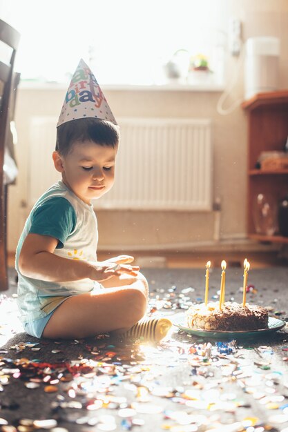 Close up photo of a small caucasian boy celebrating his birthday on the floor posing with a cake and confetti while wearing a holiday cap