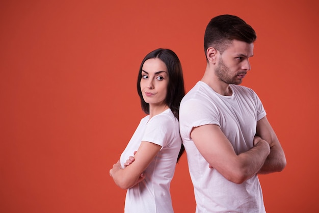 Close up photo of young angry and sad couple in white t-shirts with crossed arms and standing back to back on orange background