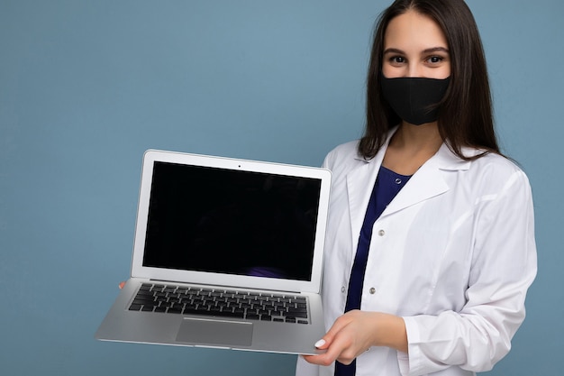 Close-up photo of Young brunet woman wearing medical white coat and black mask holding laptop