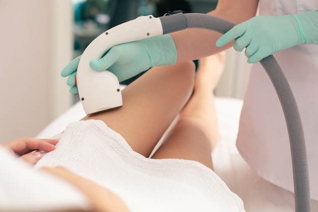 Close up photo of the young woman legs and beautician holding laser hair removal device with a special vacuum suction