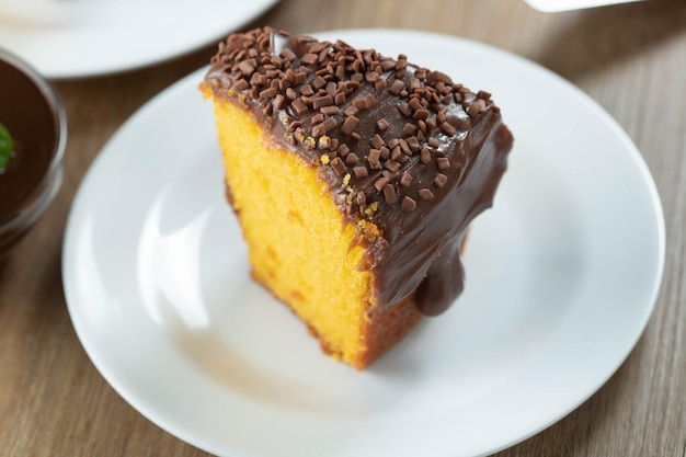 Close up piece of Brazilian carrot cake with chocolate frosting on wooden table with carrots in the background