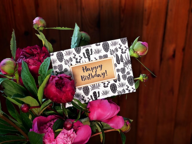 Close-up of pink peonies flowers and vase on wooden background with happy birthday card