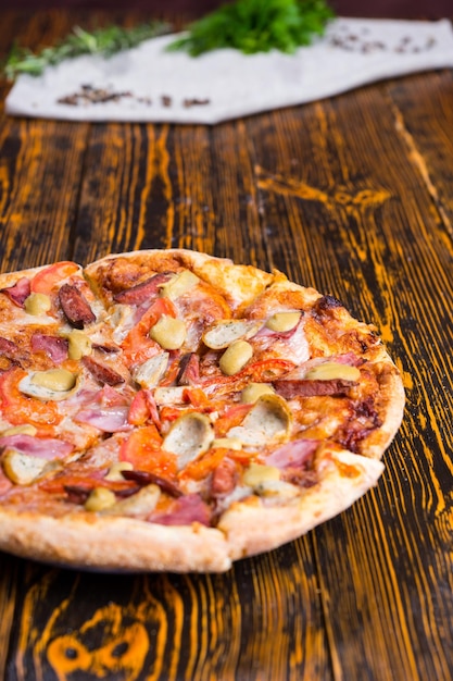 Close up of pizza with ham, near are peppers, sprig of rosemary and other spices on a napkin on wooden table