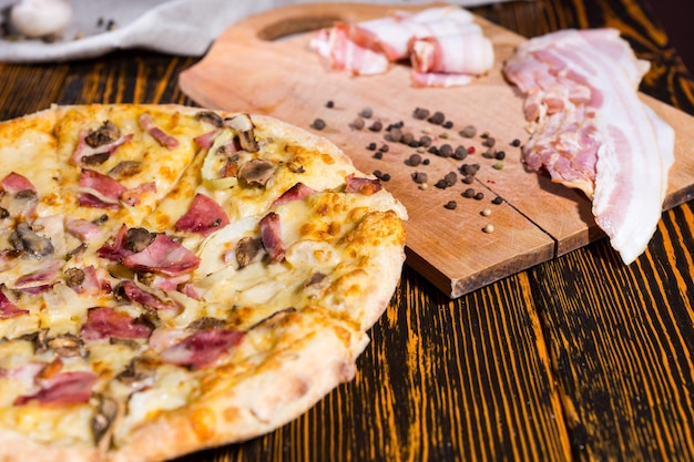 Close up of pizza with variety of toppings and cheese on wooden table, near lies cutting board with bacon and spices