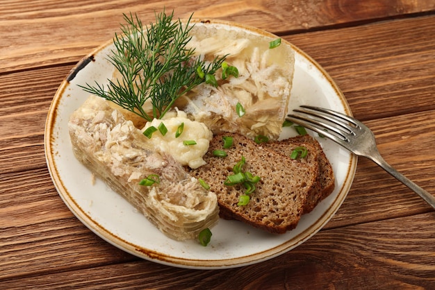 Close up portion of aspic or traditional Eastern European chicken meat jelly with slices of rye bread and horseradish dressing sauce, on brown wooden table, high angle view