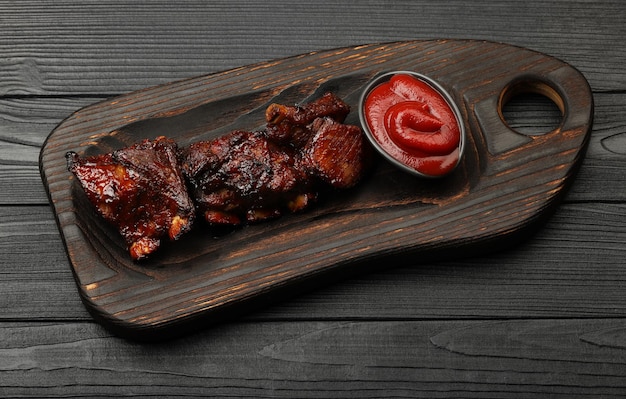 Close up portion of barbecue spare rib slices and BBQ tomato sauce served on wooden board over black table planks, high angle view, personal perspective