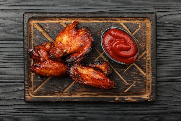 Close up portion of grilled barbecue chicken buffalo wings and BBQ tomato sauce served on wooden board over black table planks, elevated top view, directly above