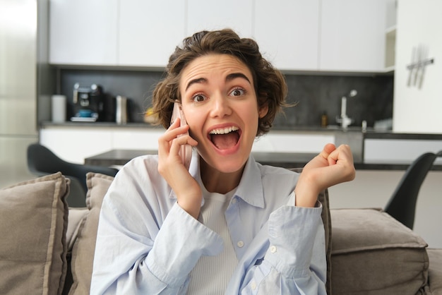 Close up portrait of amazed brunette woman jumps on sofa from excitement answers phone call hear gre