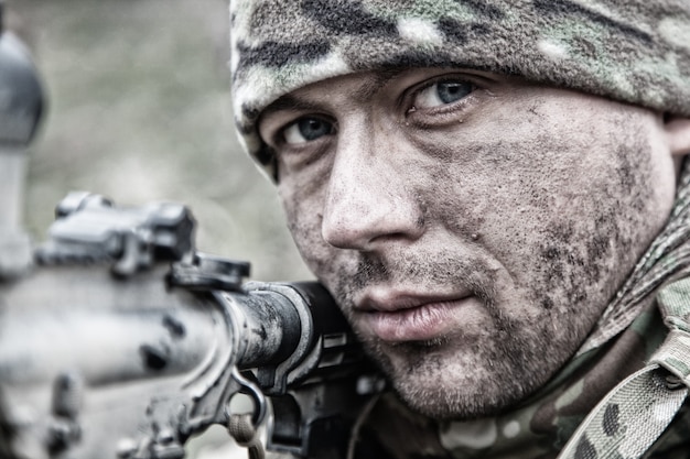 Photo close-up portrait of army infantryman, modern warfare combatant, young soldier with dirty, unshaven face, in camo bennie hat, aiming service rifle, controlling area with gunfire, looking in camera