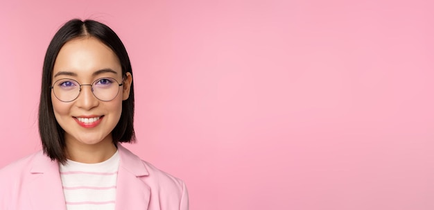 Close up portrait of asian corporate woman professional businesswoman in glasses smiling and looking confident at camera pink background