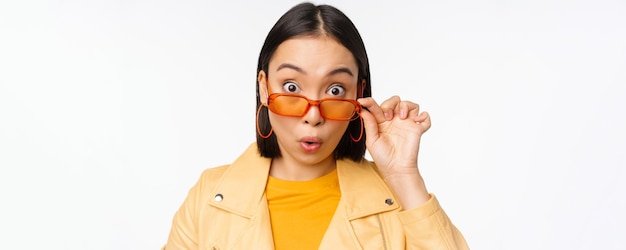 Close up portrait of asian girl looking surprised wow face takes off sunglasses and staring impressed at camera standing over white background