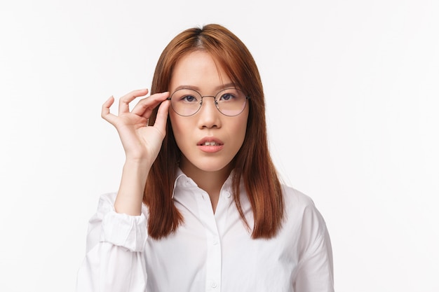 Close-up portrait of asian pretty young girl in white shirt, trying new prescribed glasses, picking eyewear at optician store, looking camera determined, seriously listening standing white