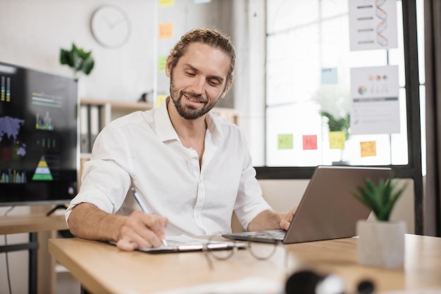 Close up portrait of bearded young male office manager in white shirt writing financial report