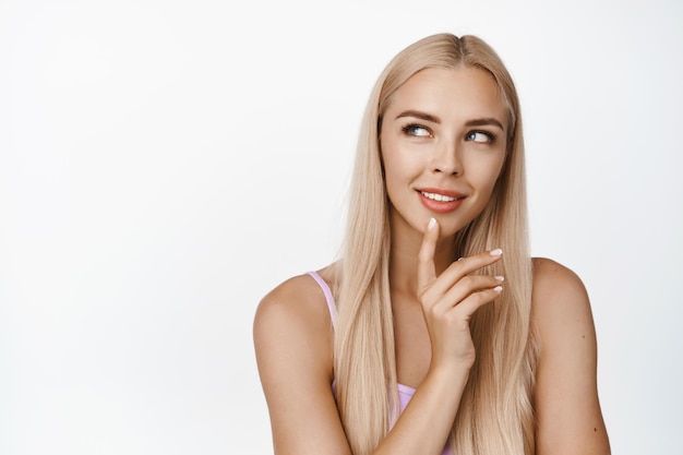 Close up portrait of beautiful blond girl with clean glowing skin perfect body looking thoughtful thinking of smth standing over white background