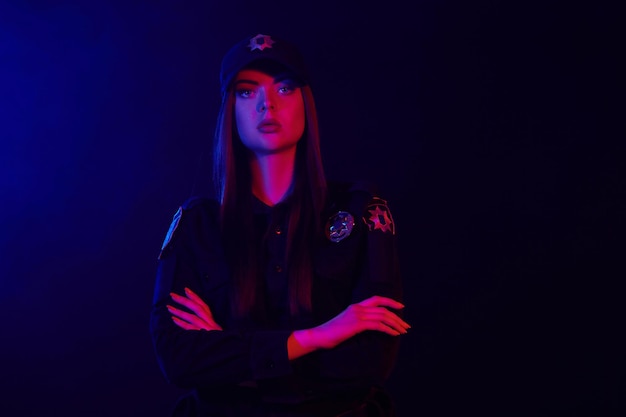 Close-up portrait of a beautiful redheaded female police officer in a uniform and a cap, with bright make-up, looking at the camera against a black background with red and blue backlighting.