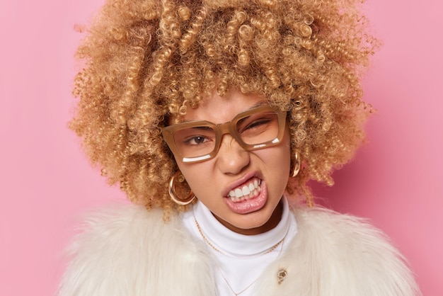 Close up portrait of beautiful woman with curly hair clenches teeth has cheeky expression has irritated gaze being dissatisfied with something dressed in winter coat isolated over pink background