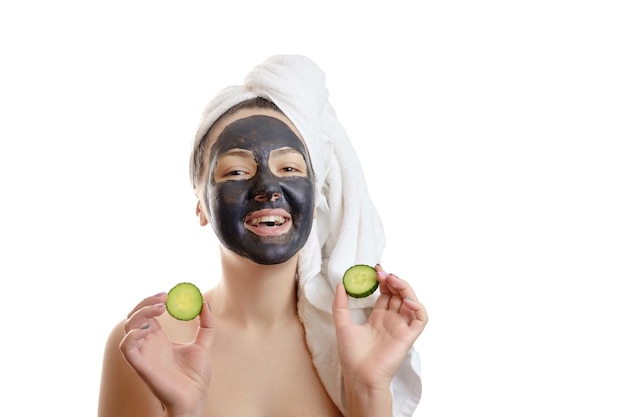 Close-up portrait beautiful woman with facial black mask and cucumber slices in her hands on white background, girl with a white towel on her head, satisfied smiles