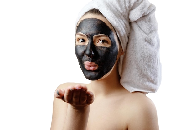 Close-up portrait beautiful woman with facial black mask on white background, girl with a white towel on her head, satisfied and happy smile, air kiss