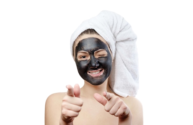 Close-up portrait beautiful woman with facial black mask on white background, girl with a white towel on her head, satisfied and happy smile, pointing hands at the camera