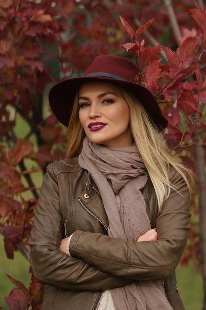 Close-up portrait of beautiful young blonde girl in burgundy hat with feather and scarf on background of sunny autumn park