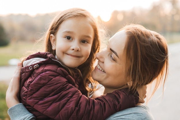 Close up portrait of beautiful young mother embracing her little girl which is  smiling outdoor in the park.