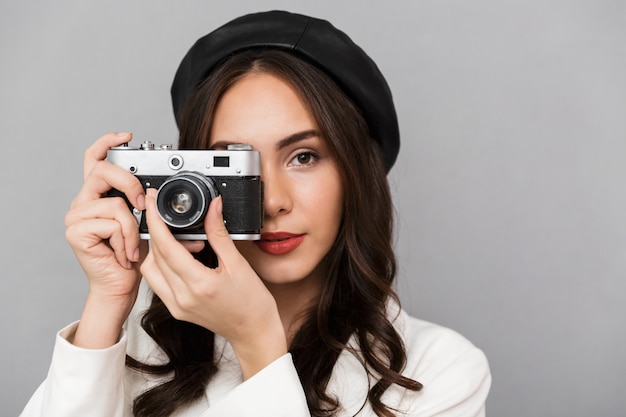 Close up portrait of a beautiful young woman dressed in jacket over gray background, taking picture with photo camera