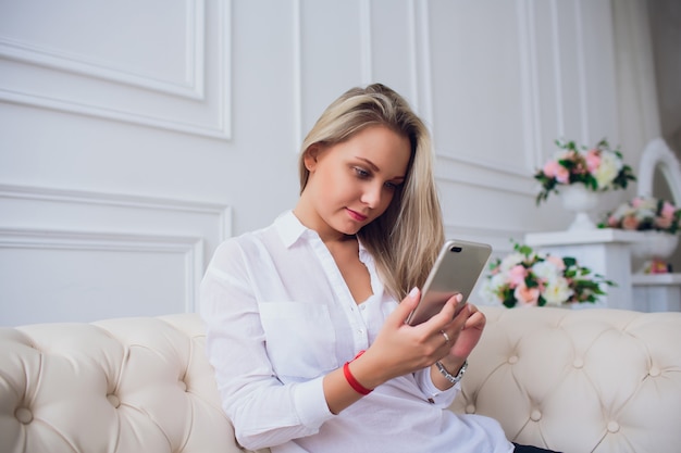 Close-up portrait of blond female sitting back to back on sofa and using her smart phone. Beautiful woman touching the screen and making call.