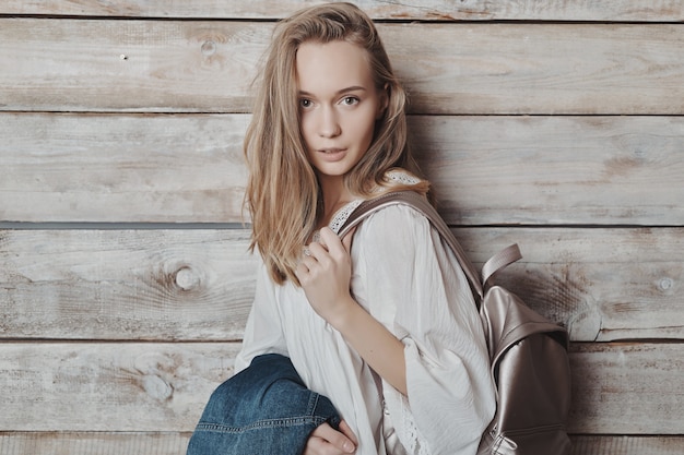 Close up portrait blonde young girl with silver backpack over wood wall