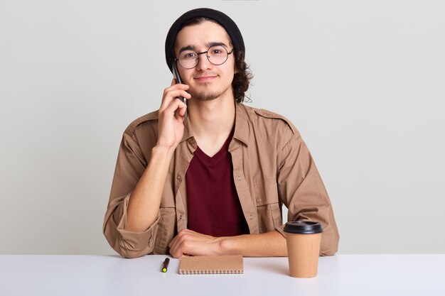 Close up portrait of busy creative man calls business partner to discuss future meeting, has new idea, freelancer writing something in his notebook and drinking coffee, isolated over white studio wall