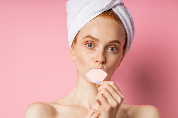 Close up portrait of caucasian ginger woman with freckles, clean skin, bare shoulders, covering mouth with cosmetic sponge, having fun after shower, wearing towel on head isolated pink background.