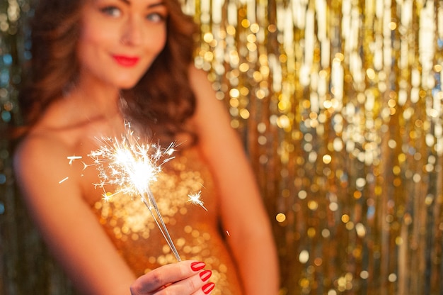 Close-up portrait of Caucasian woman in gold dress holding sparkler at party on bright gold background, selective focus