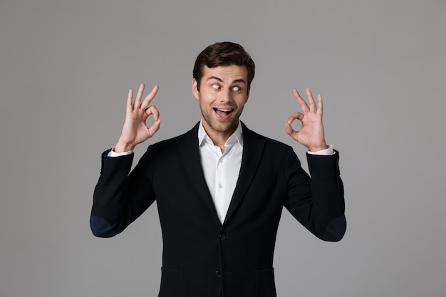 Close up portrait of a cheerful young businessman dressed in suit isolated over gray wall, showing ok gesture