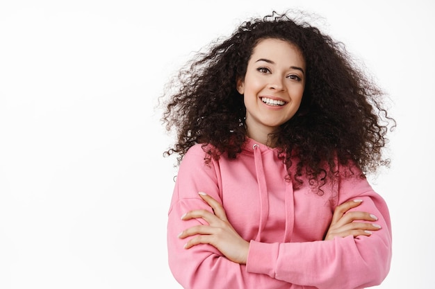 Close up portrait of confident brunette woman in casual hoodie, cross arms on chest, smiling and looking happy, standing against white background