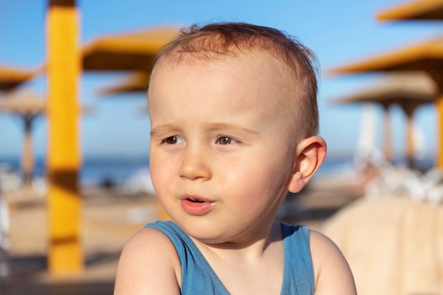 Close up portrait of a cute adorable toddler boy looking aside surprised and charmed.Llittle funny baby child is sitting on the beach of the tropical resort in summer noon.