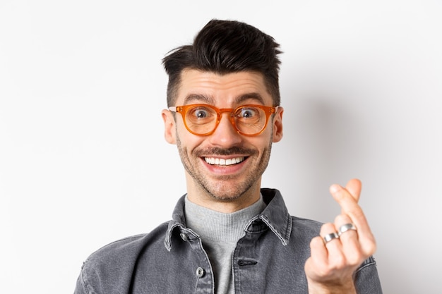 Close up portrait of cute guy with moustache wearing glasses, showing hand heart sign, smiling happy, standing on white background.