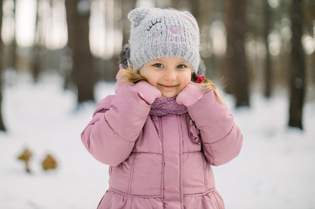 Close up portrait of cute pretty little girl in pink jacket and gray cap, smiling at camera on beautiful winter snow background outdoors in the forest