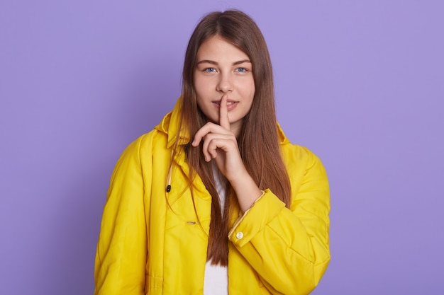 Close up portrait of cute winsome pretty woman holding forefinger near lips isolated on lilac background, has confident facial expression, wearing bright yellow jacket, keeps secret information.