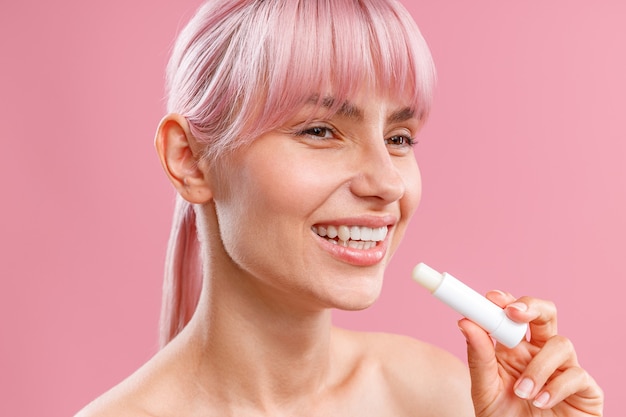 Photo close up portrait of cute young woman with pink hair smiling aside holding lip balm near her lips