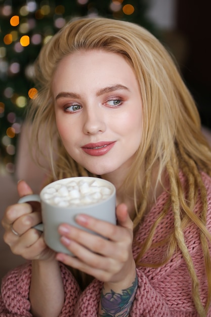 Close-up portrait of dreaming happy girl with dreadlocks holding a mug of cocoa with marshmallows