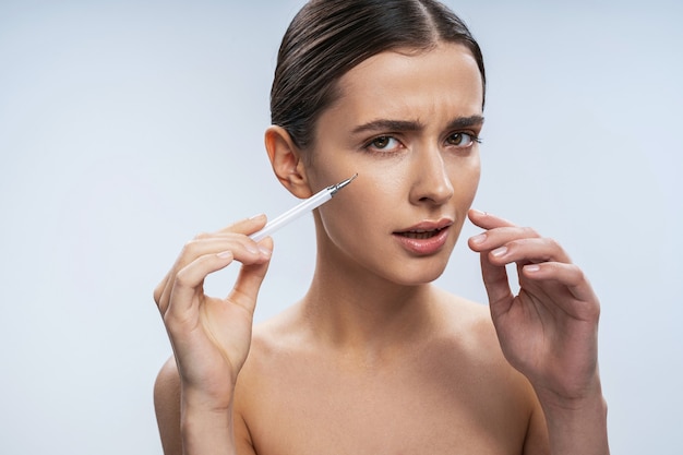 Close up portrait of elegant Caucasian female doing hyaluronic acid shot under her eyes isolated on white background